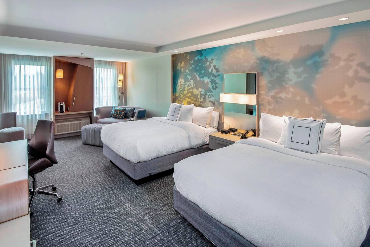 A hotel room with two beds, a desk, a chair, and a large window with curtains.