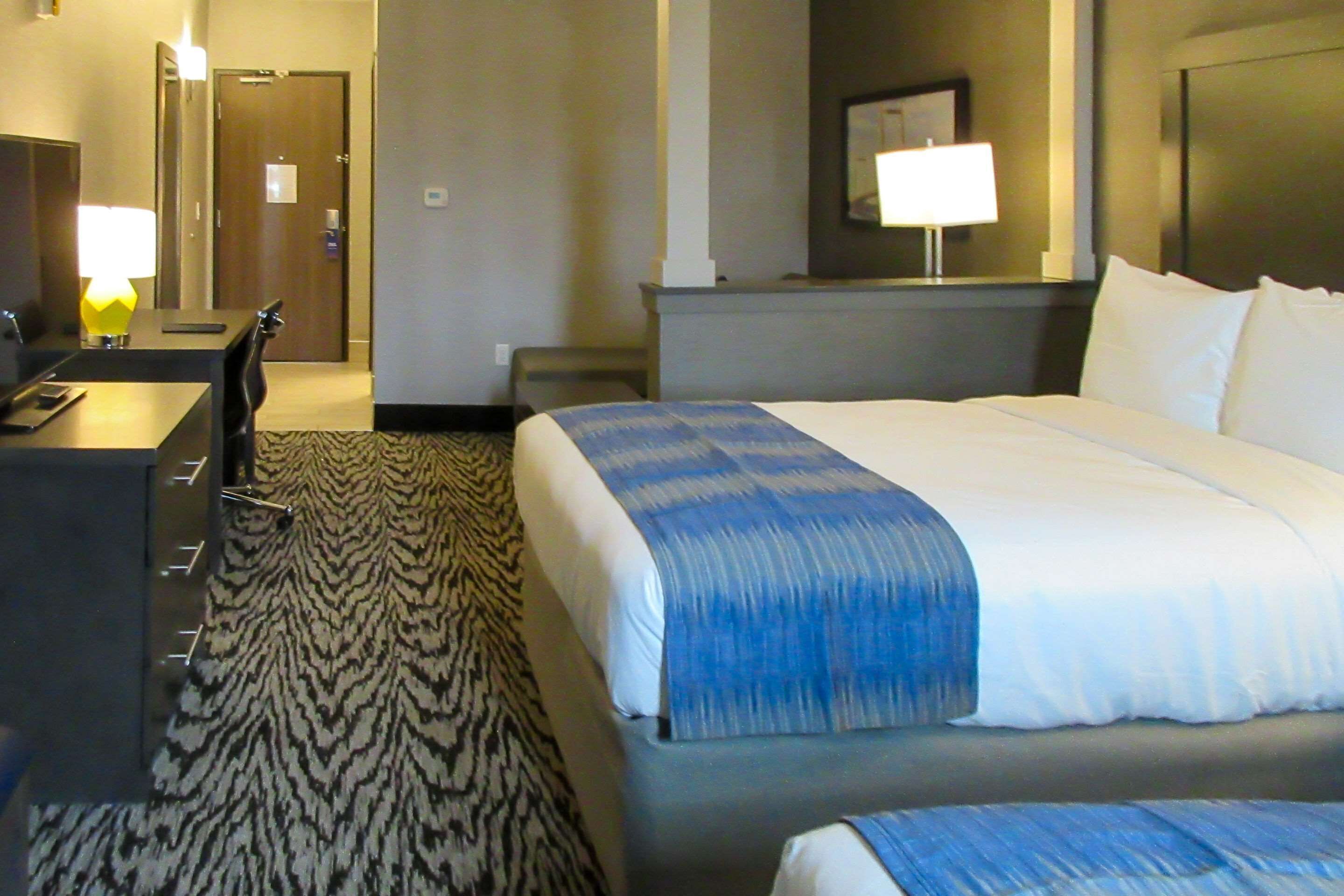 A hotel room with a bed, a desk with a lamp, a chair, and a carpet.