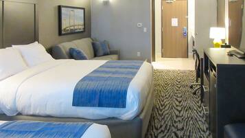 Habitación de hotel con dos camas, un sofá, un escritorio con lámpara y una puerta que da a otra habitación.