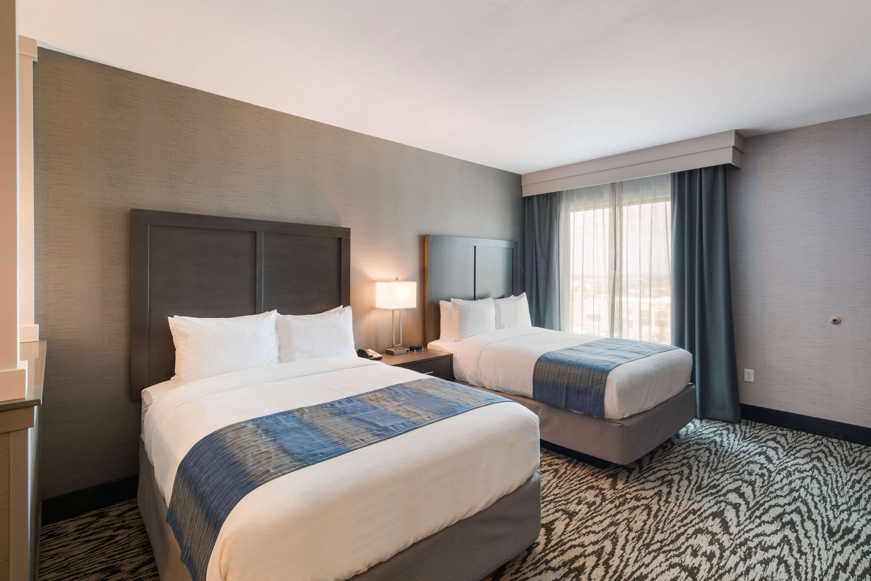 Una habitación de hotel con dos camas, una mesita de noche con lámpara y un ventanal con cortinas.
