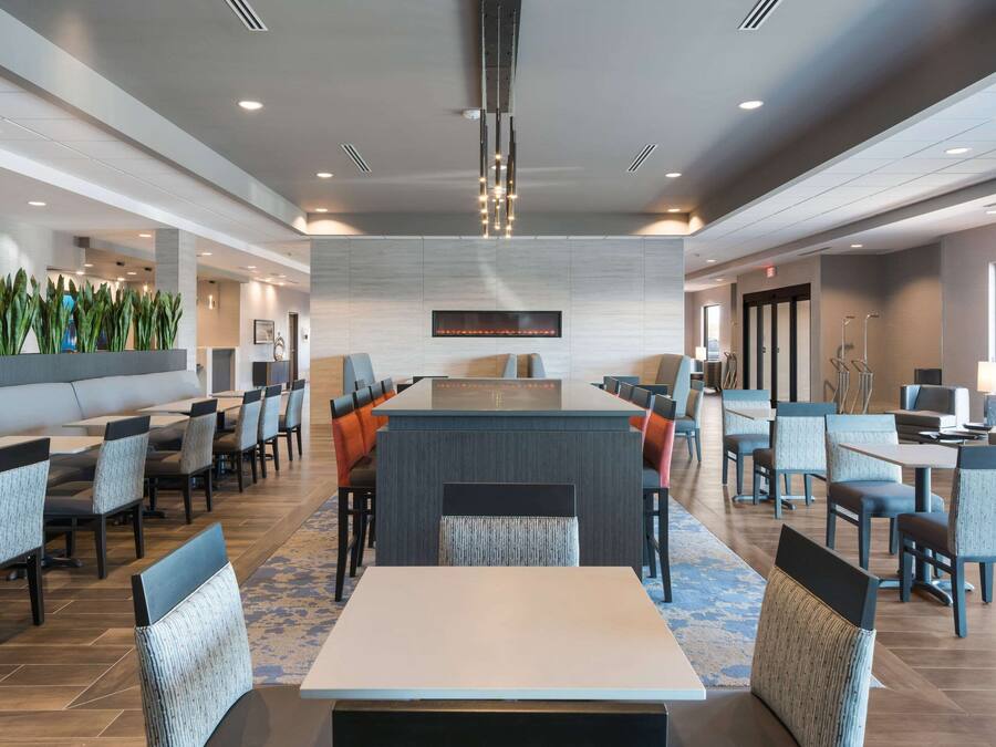A modern hotel lobby with a central seating area, multiple tables, and chairs, and a large wall-mounted TV.