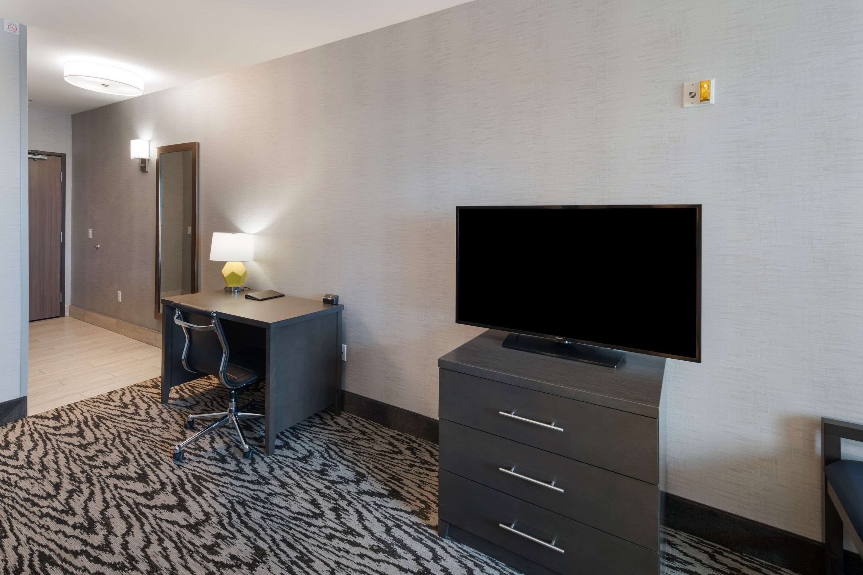 Ein Hotelzimmer mit Flachbildfernseher, Schreibtisch mit Lampe, Stuhl, dunklem Kommodenstück und einem gemusterten Teppich.