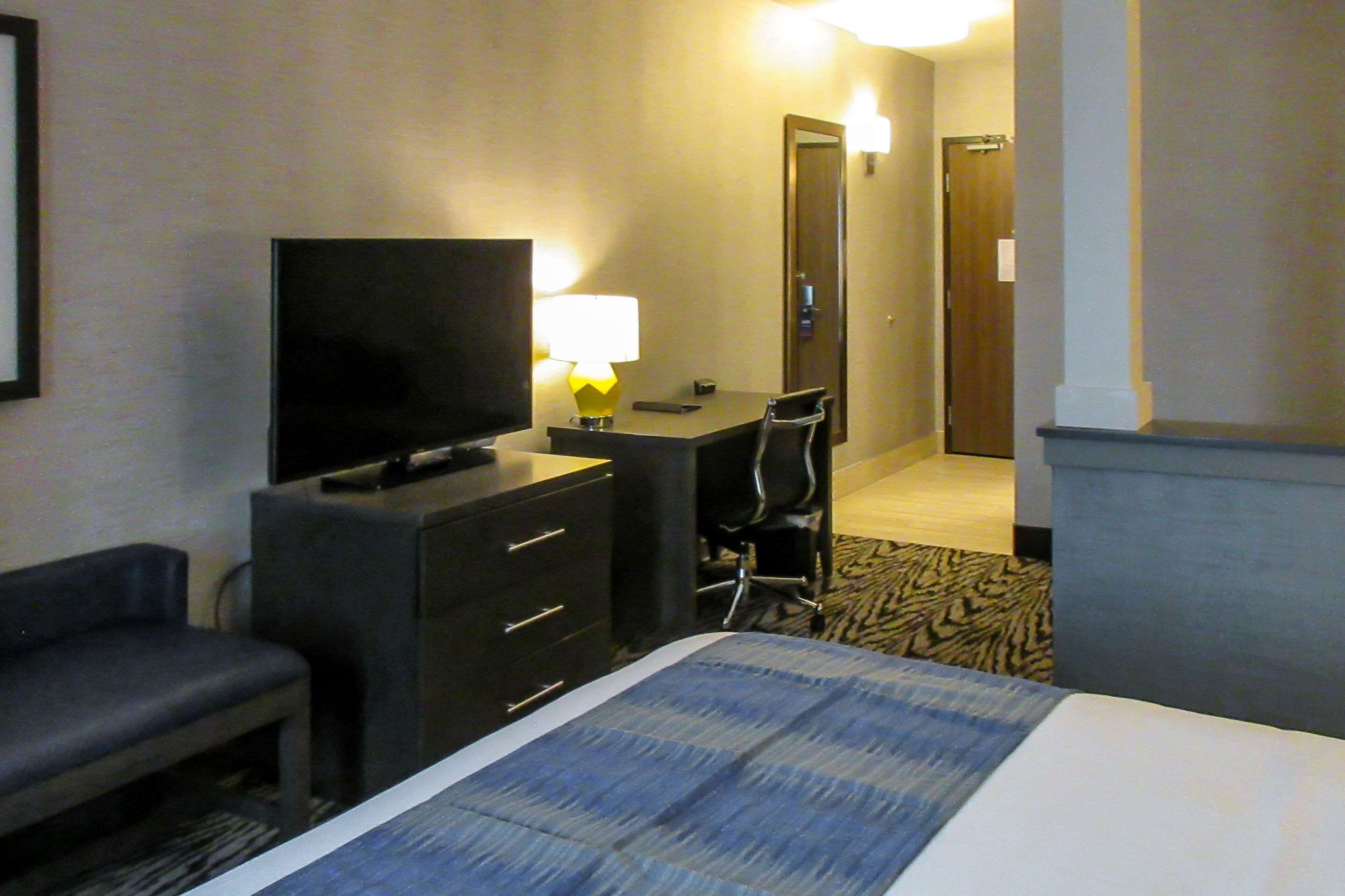 A hotel room with a bed, a TV, a desk with a lamp, a chair, and a small bench.