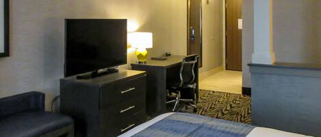 Una habitación de hotel con cama, televisión, escritorio con lámpara, silla y una pequeña banca.