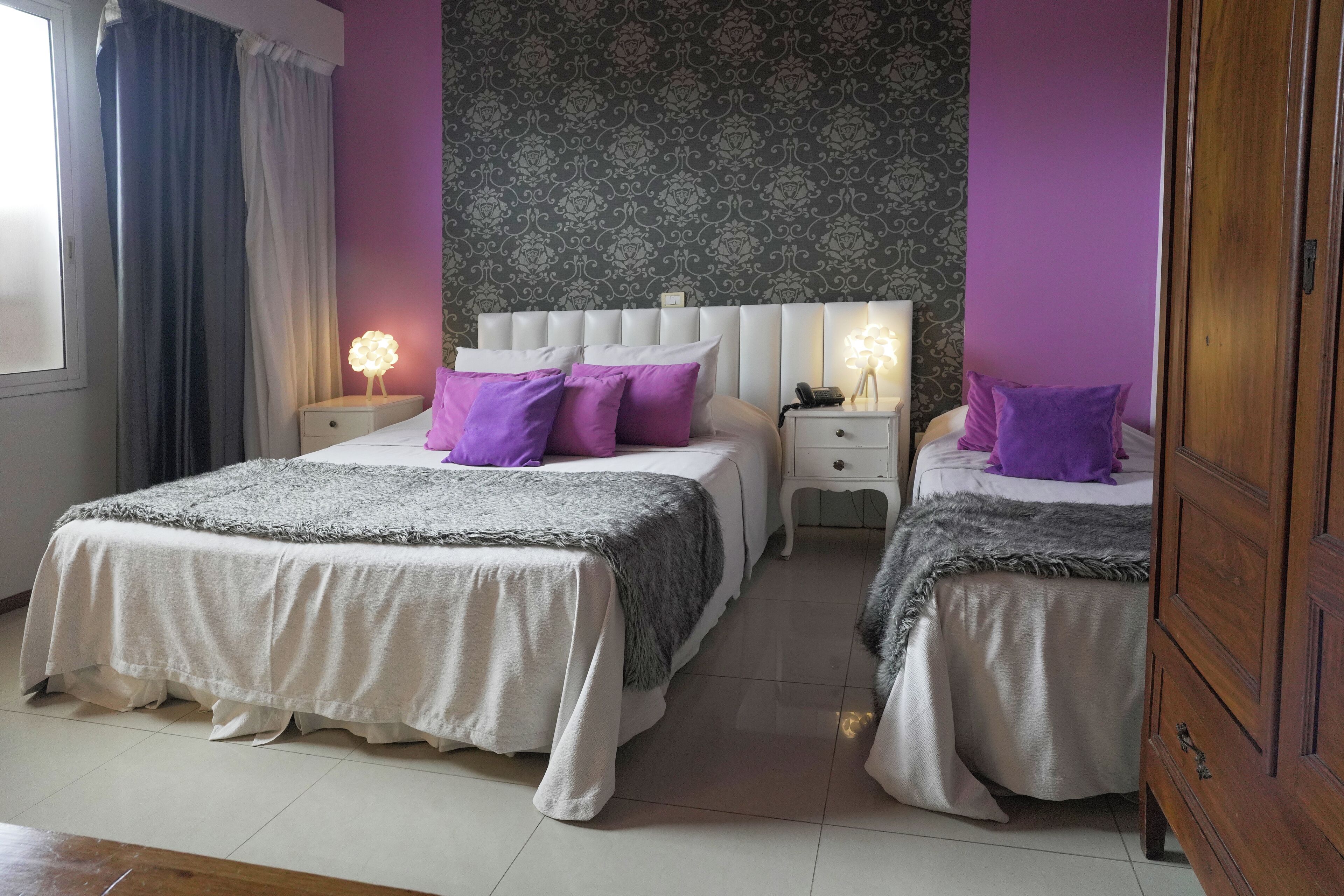 A double room with two beds, each with a white headboard and purple pillows. There are bedside tables with lamps, a large window with curtains, and a patterned wallpaper on the wall.