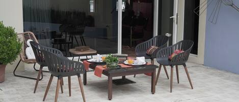 En udendørs terrasse med et bord dækket til to personer, omgivet af fire stole og en potteplante ved siden af.