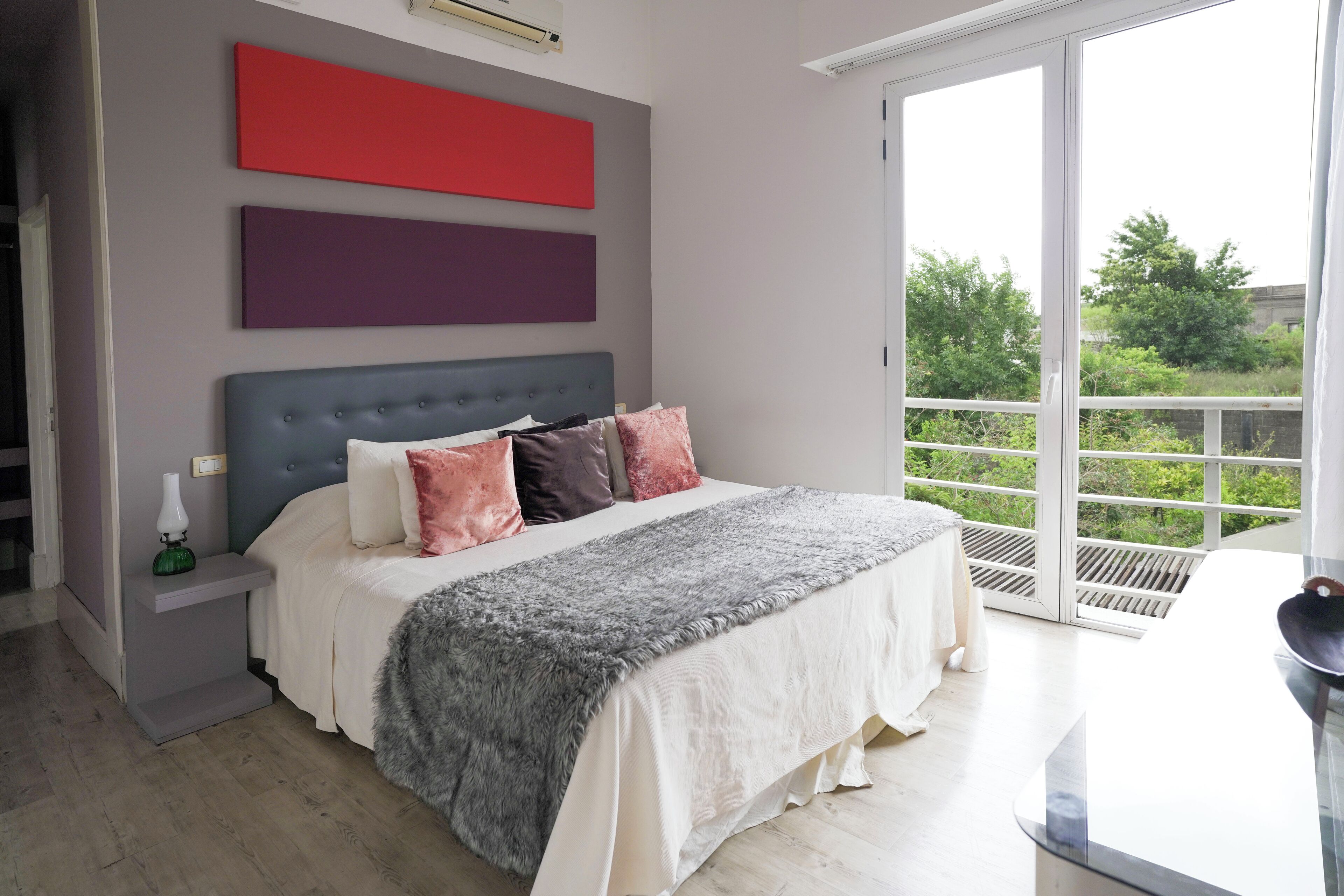 A bedroom with a bed, a nightstand, a window with a view of greenery, and a red and purple wall art piece.