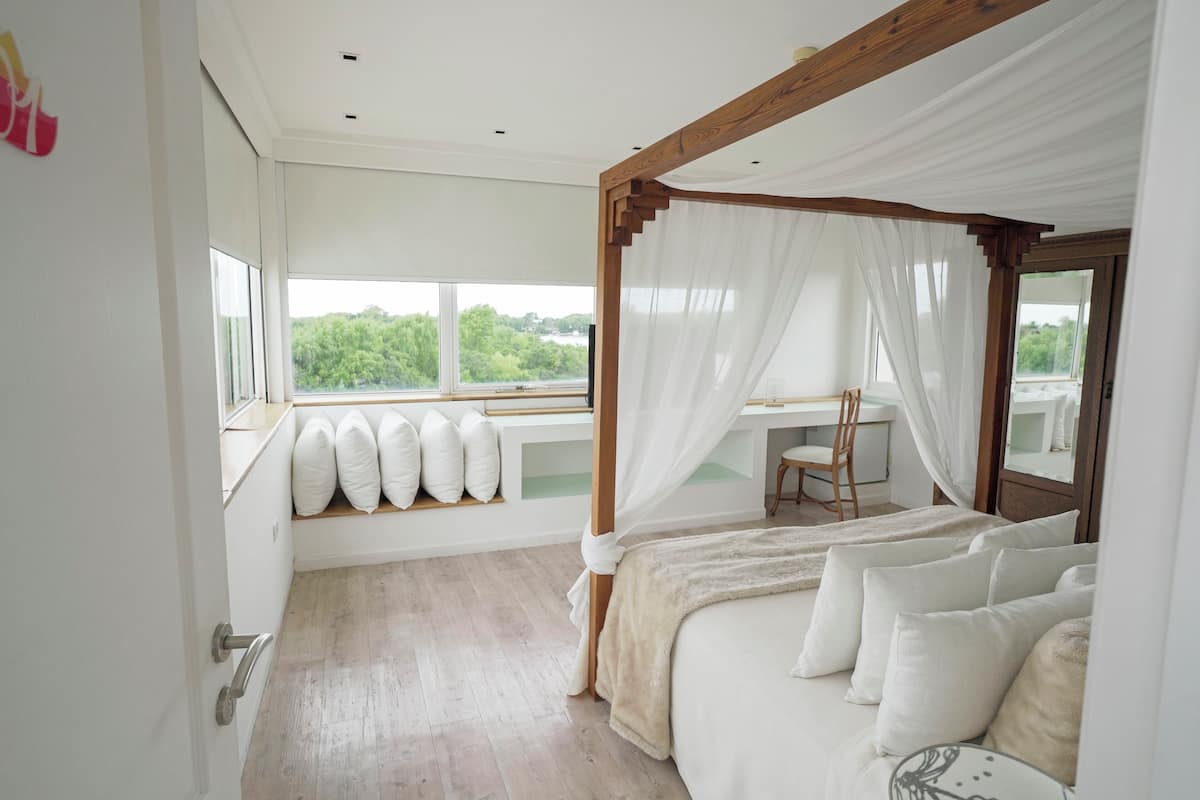 Une chambre à coucher avec un lit à baldaquin, un bureau avec une chaise et une vue sur la verdure grâce à de grandes fenêtres.