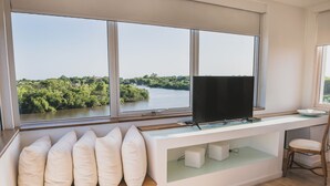 A modern room with a large window overlooking a river and greenery, a flat-screen TV on a white console, and a wooden bench with cushions. - Hotel Boutique Los Muelles (Carmelo)