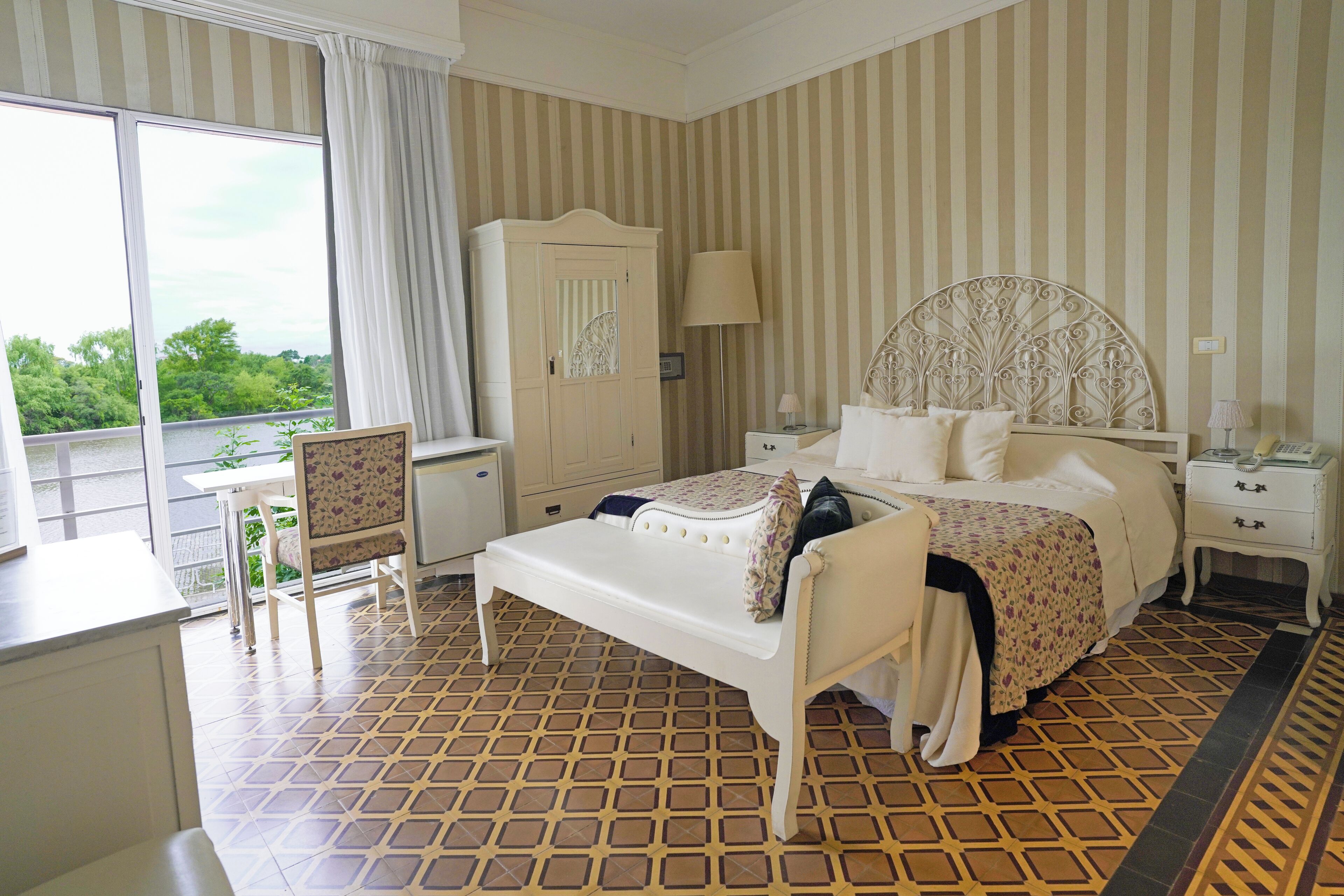A hotel room with a bed, a chair, a desk, a wardrobe, and a view of greenery outside.