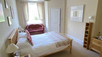 A bedroom with a bed, a pink sofa, a bedside table with a lamp, a mirror, and a shelf with decorative items.