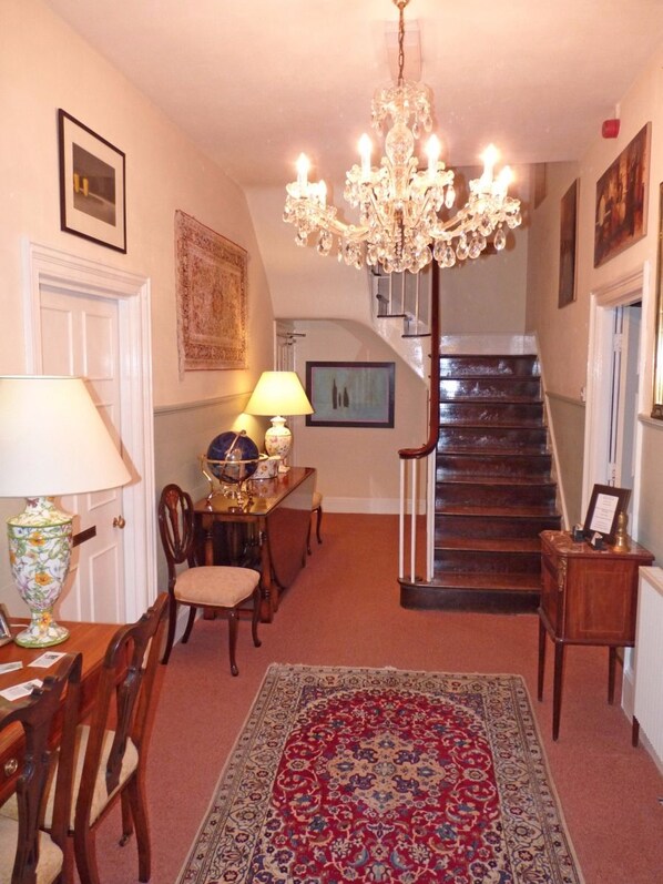 Una escalera que lleva al segundo piso, un candelabro, un área de comedor con mesa y sillas, y una alfombra estampada.