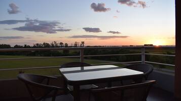 Un balcón con mesa y sillas, con vista a una puesta de sol sobre un campo.