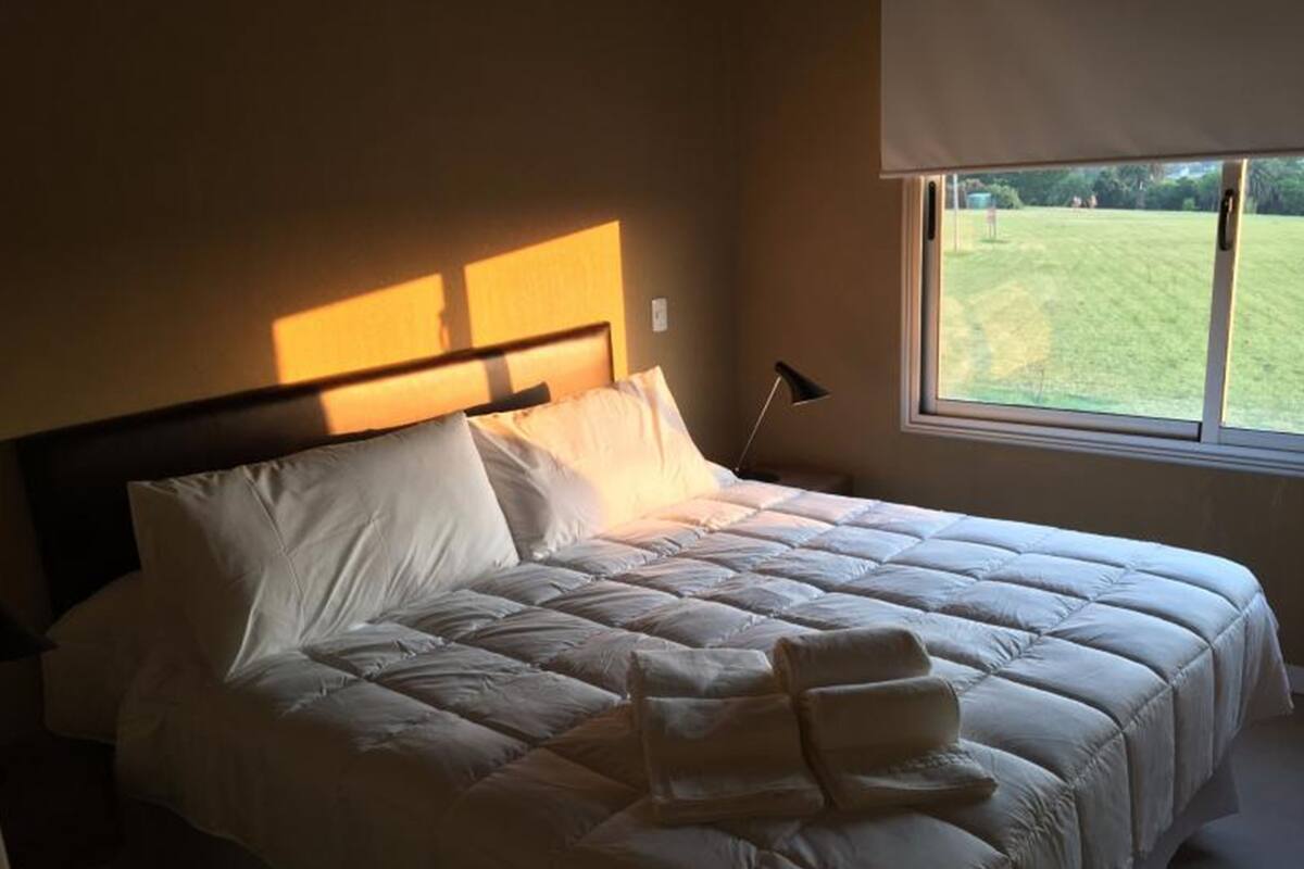 A neatly made bed with a headboard, a bedside lamp, and a window with a view of a grassy area.