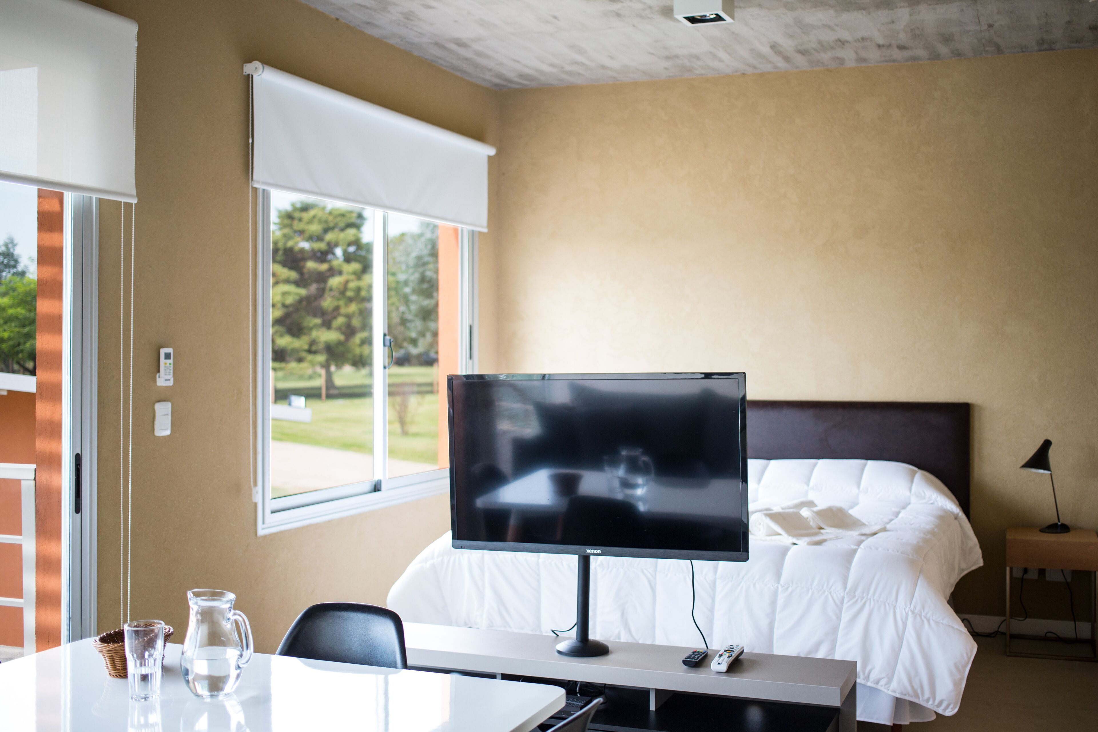 Hotellihuone, jossa on sänky, työpöytä tietokonenäytöllä, ikkuna, josta avautuu näkymä vehreään maisemaan, sekä yöpöydällä oleva lamppu.