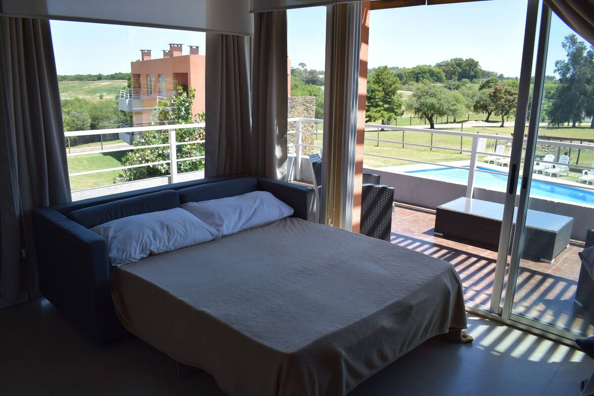 A bedroom with a large bed, a balcony with a pool, and a view of the surrounding landscape.