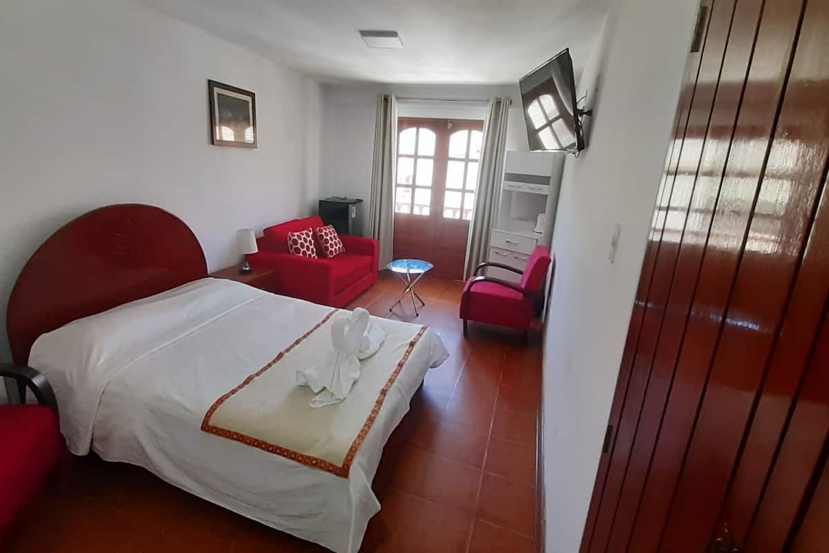 A hotel room with a bed, a red sofa, a small table, and a TV mounted on the wall.