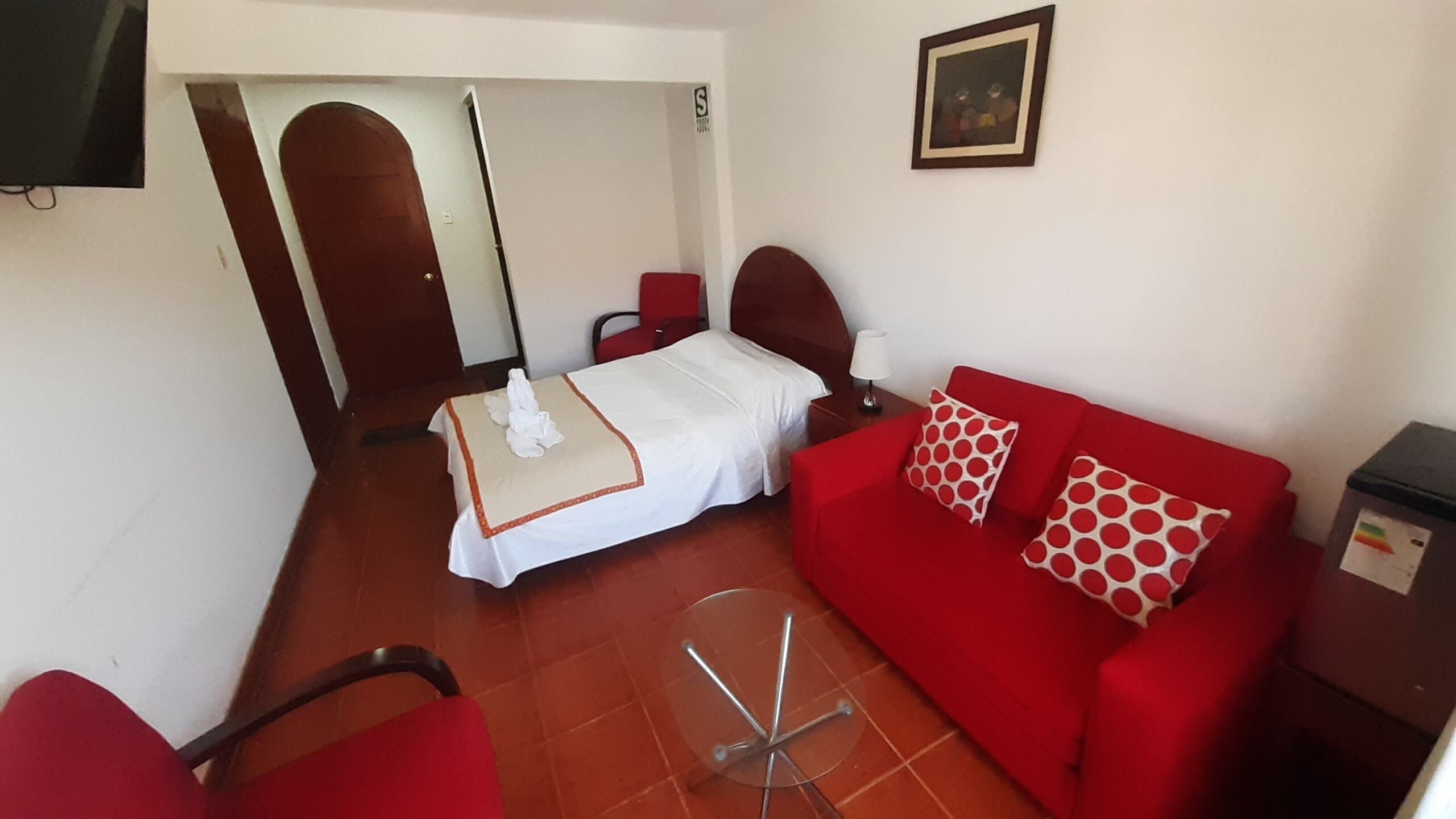 A hotel room with a bed, a red sofa with patterned cushions, a small table, and a TV mounted on the wall.