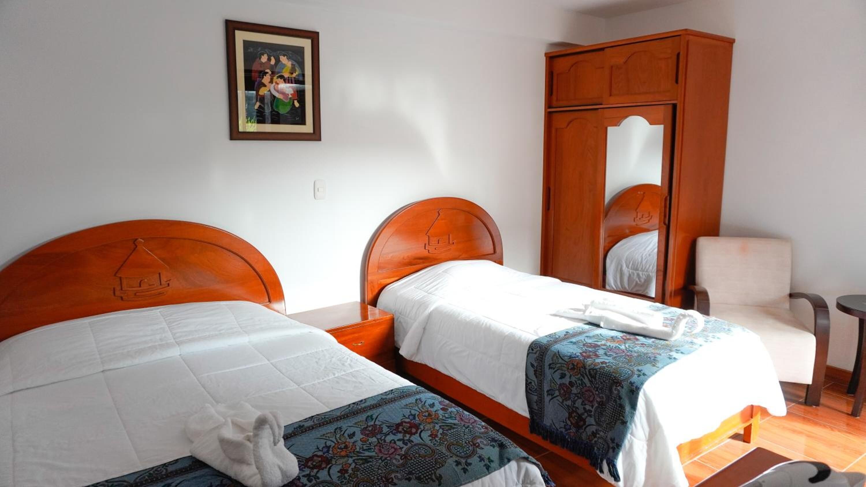 Quarto de hotel com duas camas, um guarda-roupa de madeira, uma cadeira e um quadro na parede.