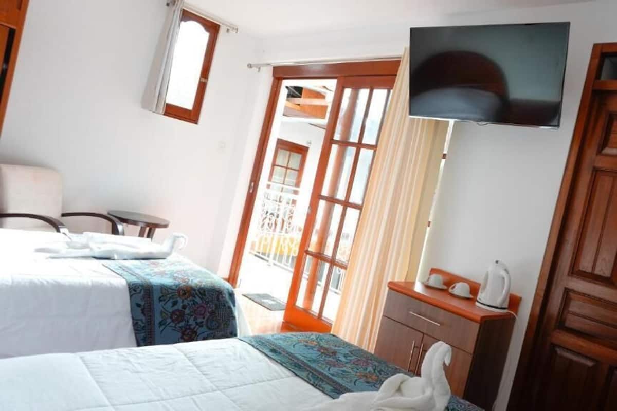 A hotel room with two beds, a TV mounted on the wall, a wooden door, and a balcony with curtains.