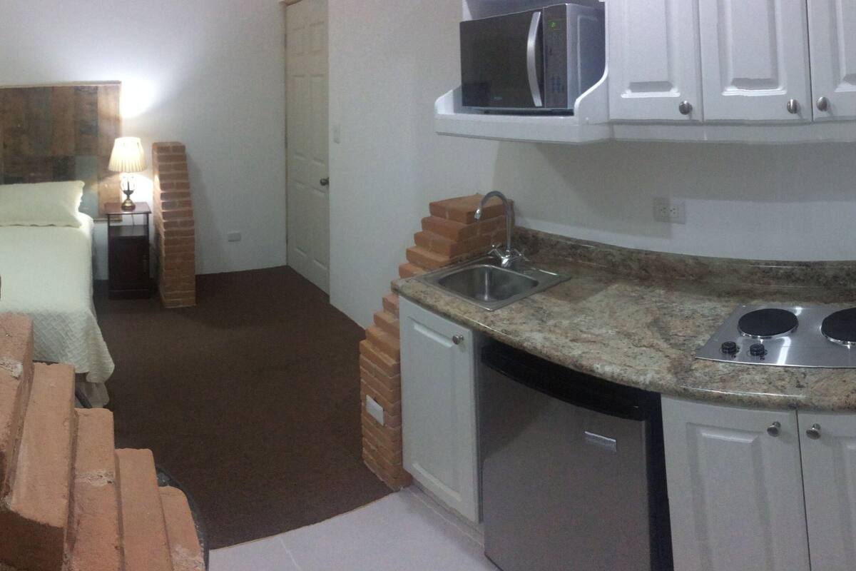 Cocina compacta con microondas, estufa y pileta. Se observa una pared de ladrillo en un lateral y una cama con mesita de noche al fondo.