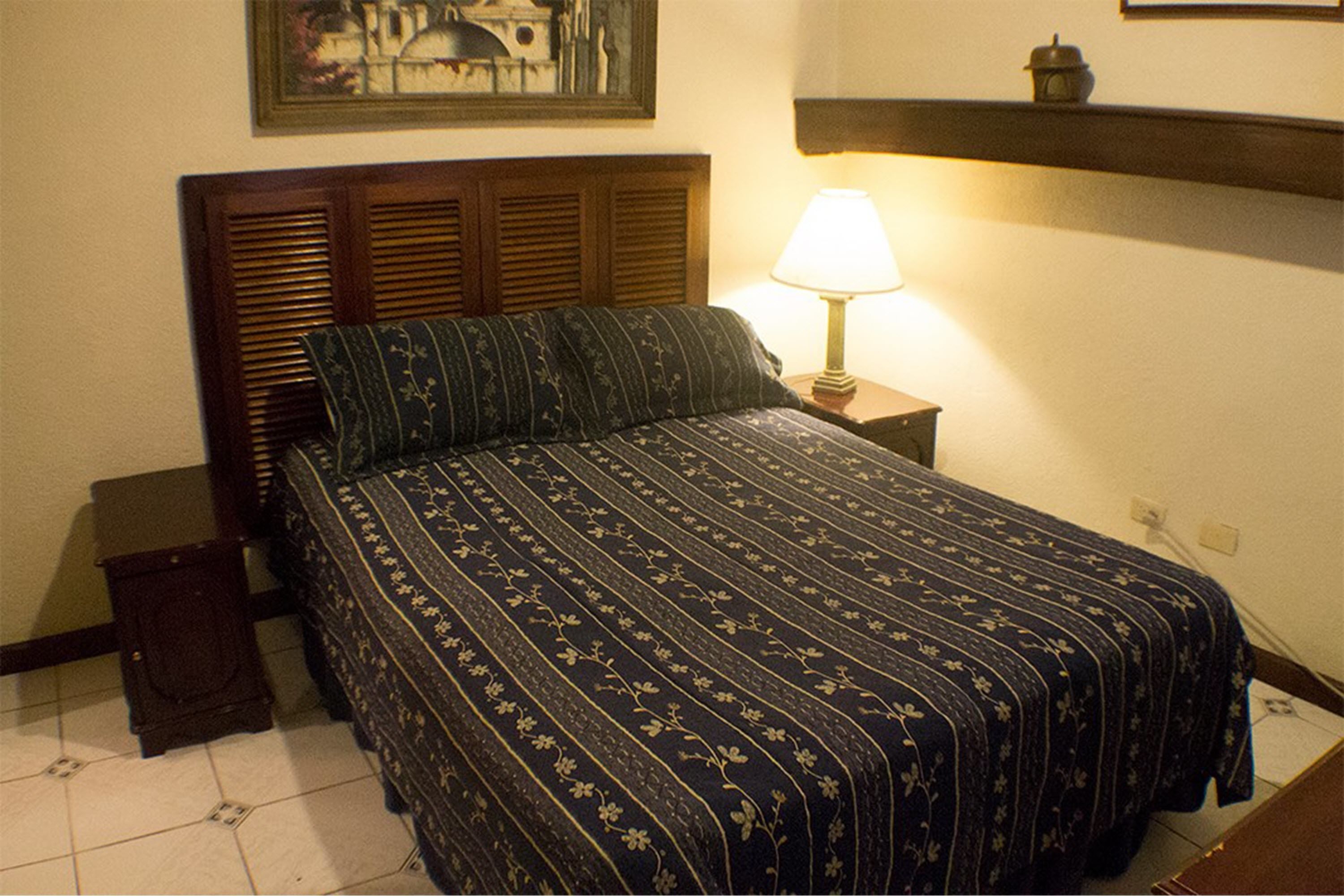 Una cama bien hecha con una colcha de estampado floral y un cabecero de madera. Hay una mesita de noche con una lámpara y una pequeña superficie auxiliar.