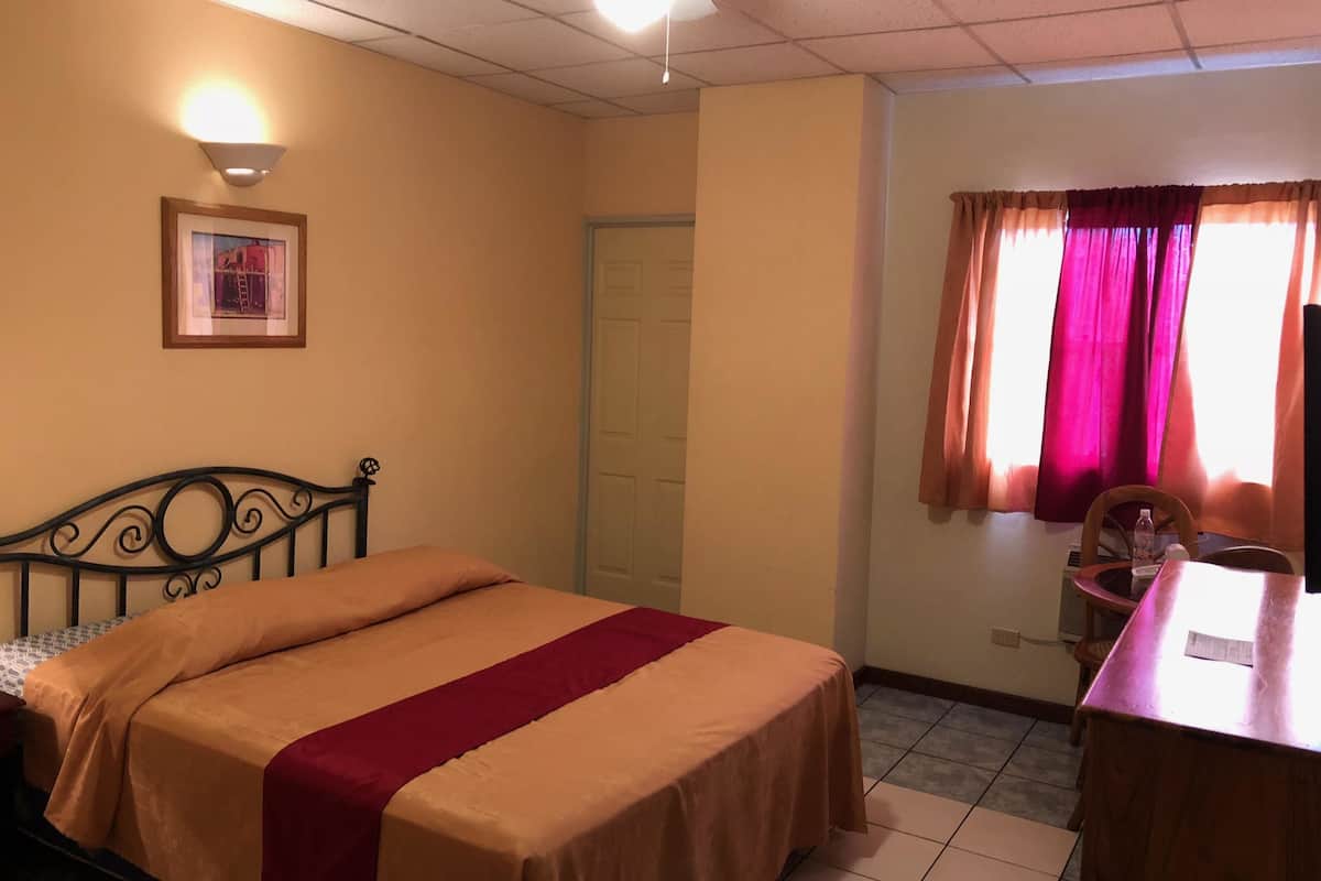 Ein Hotelzimmer mit Bett, Schreibtisch, Sessel, einem gerahmten Bild an der Wand und einem Fenster mit Vorhängen.