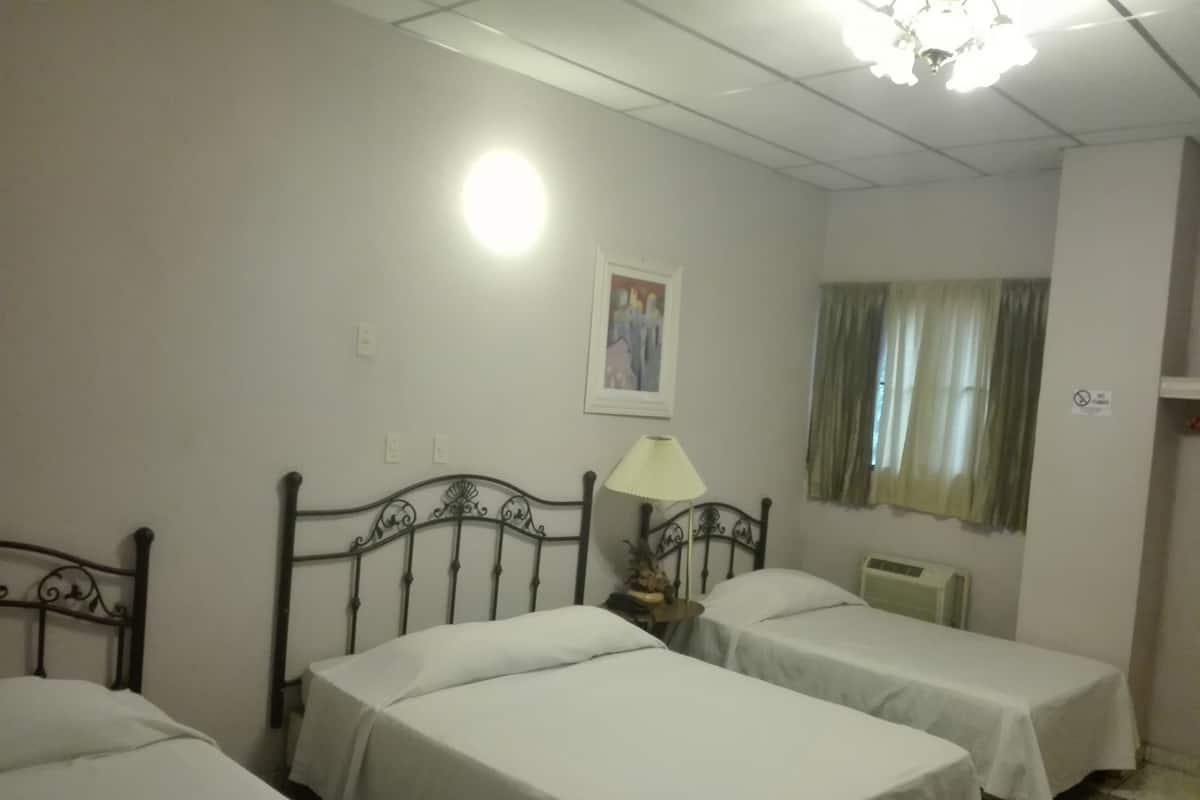 Ein Hotelzimmer mit zwei Einzelbetten, einem Nachttisch mit Lampe, einem Heizkörper, einem Fenster mit Vorhängen und einer Wandleuchte.