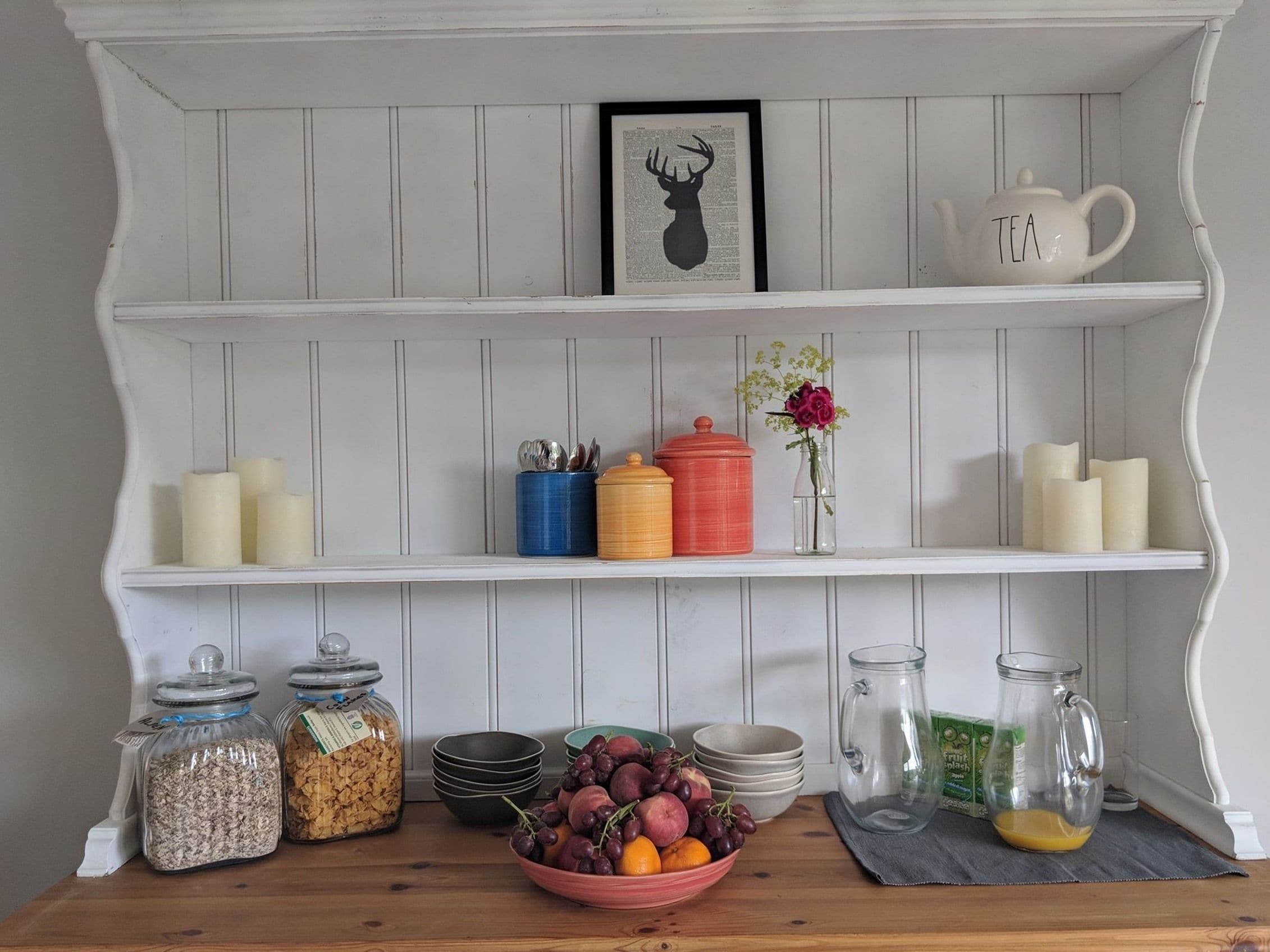 A kitchen with white wooden shelves, jars of grains, a bowl of fruit, and a tea pot.