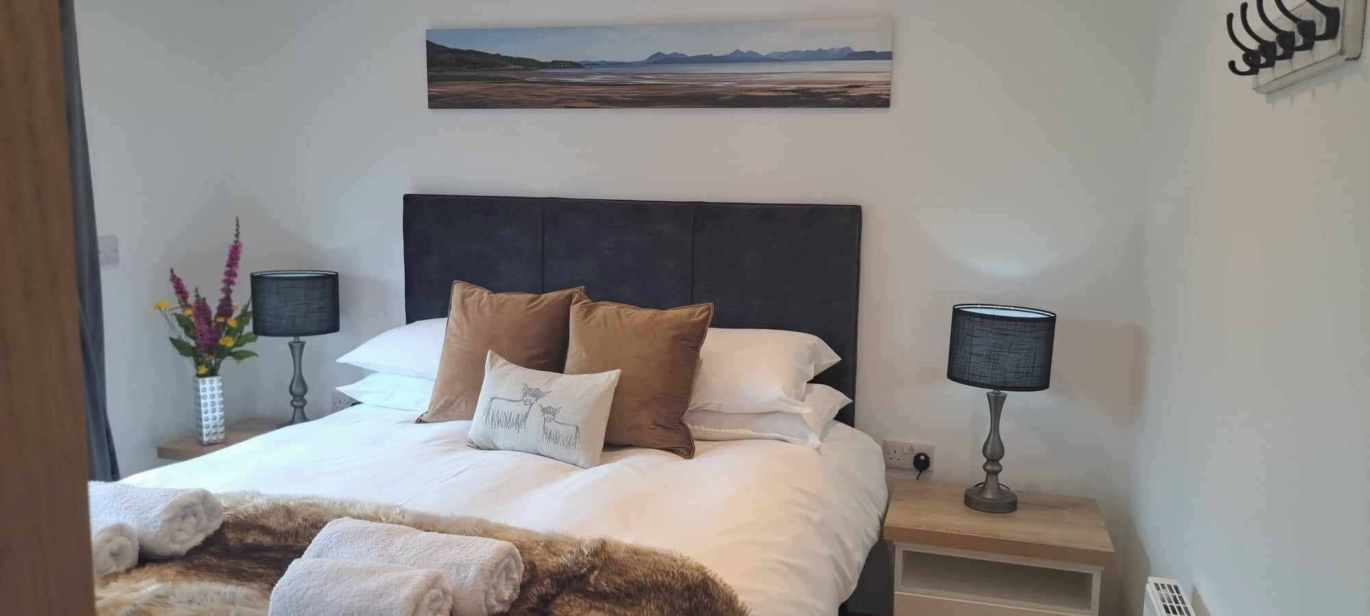 A neatly made bed with a headboard, two bedside lamps, a nightstand, and a painting on the wall.