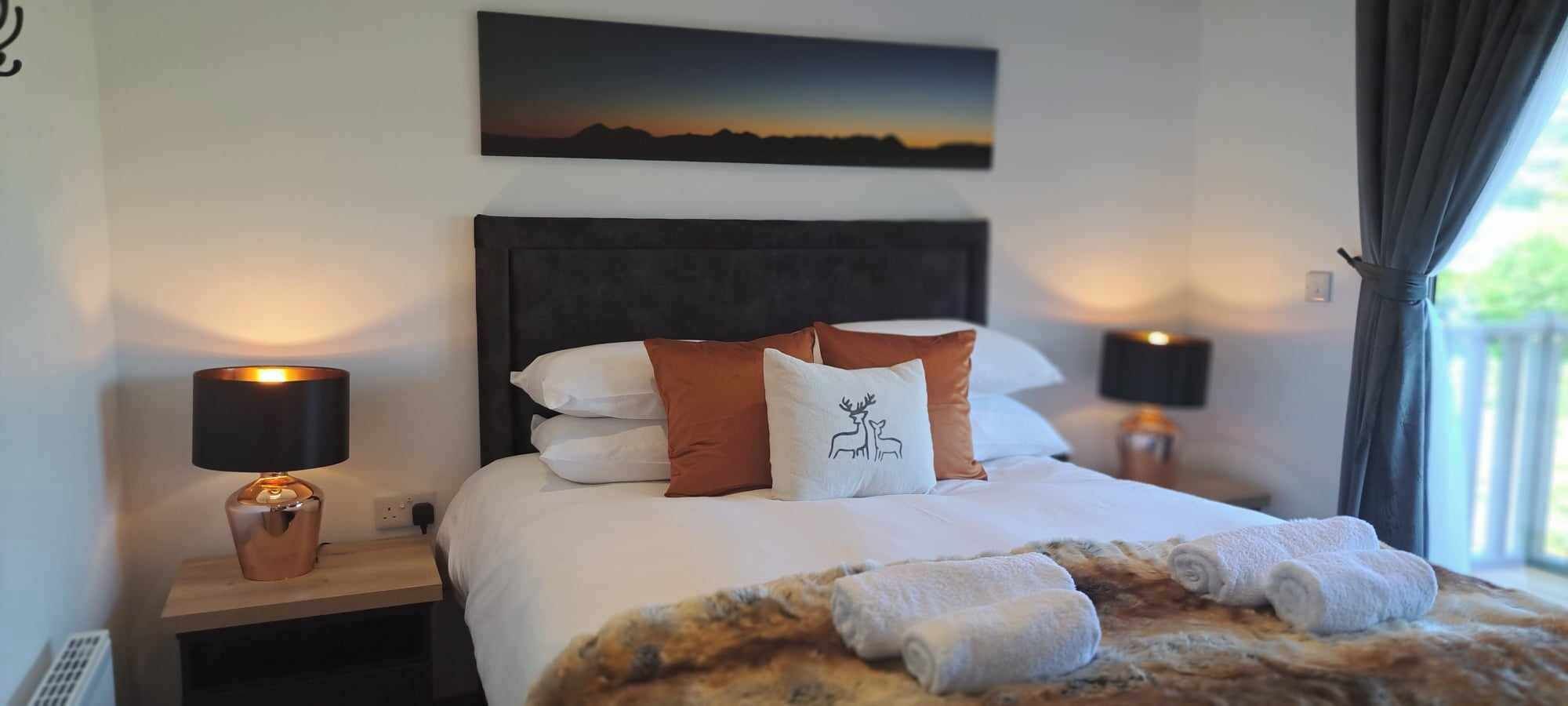 Un lit bien fait, avec du linge de lit blanc, un coussin décoratif orné d’un motif de cerf et une couverture à motifs imitant la fourrure. On y trouve deux lampes de chevet avec des abat-jour noirs, et un tableau représentant un paysage de montagne au-dessus du lit.