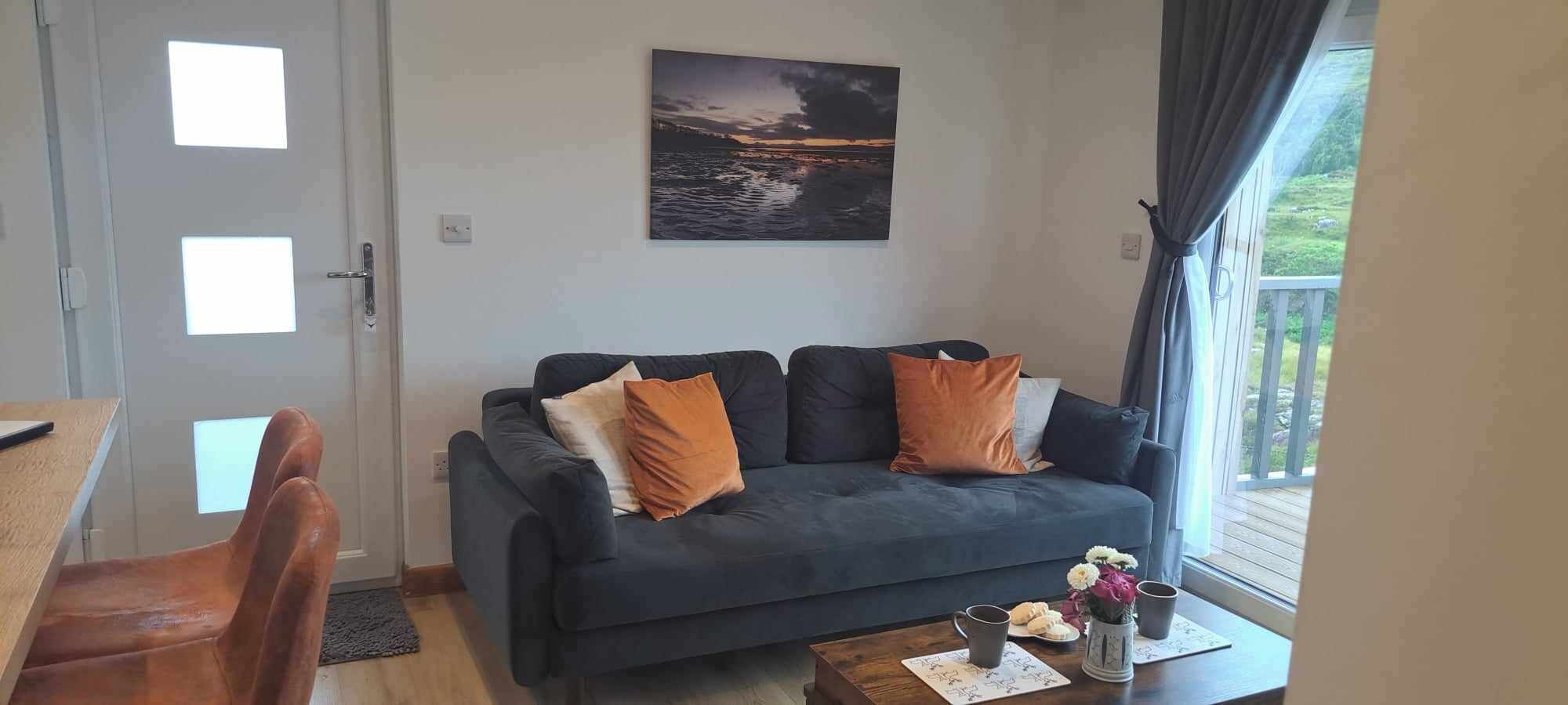 A living room with a dark blue sofa, two orange cushions, a coffee table with a vase of flowers, and a painting of a sunset over water on the wall.