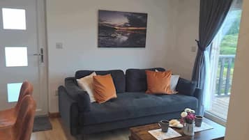 A living room with a dark blue sofa, two orange cushions, a coffee table with a vase of flowers, and a painting of a sunset over water on the wall.