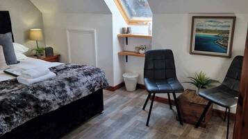 Un dormitorio con una cama, dos sillas, una mesita, un cuadro y un tragaluz.