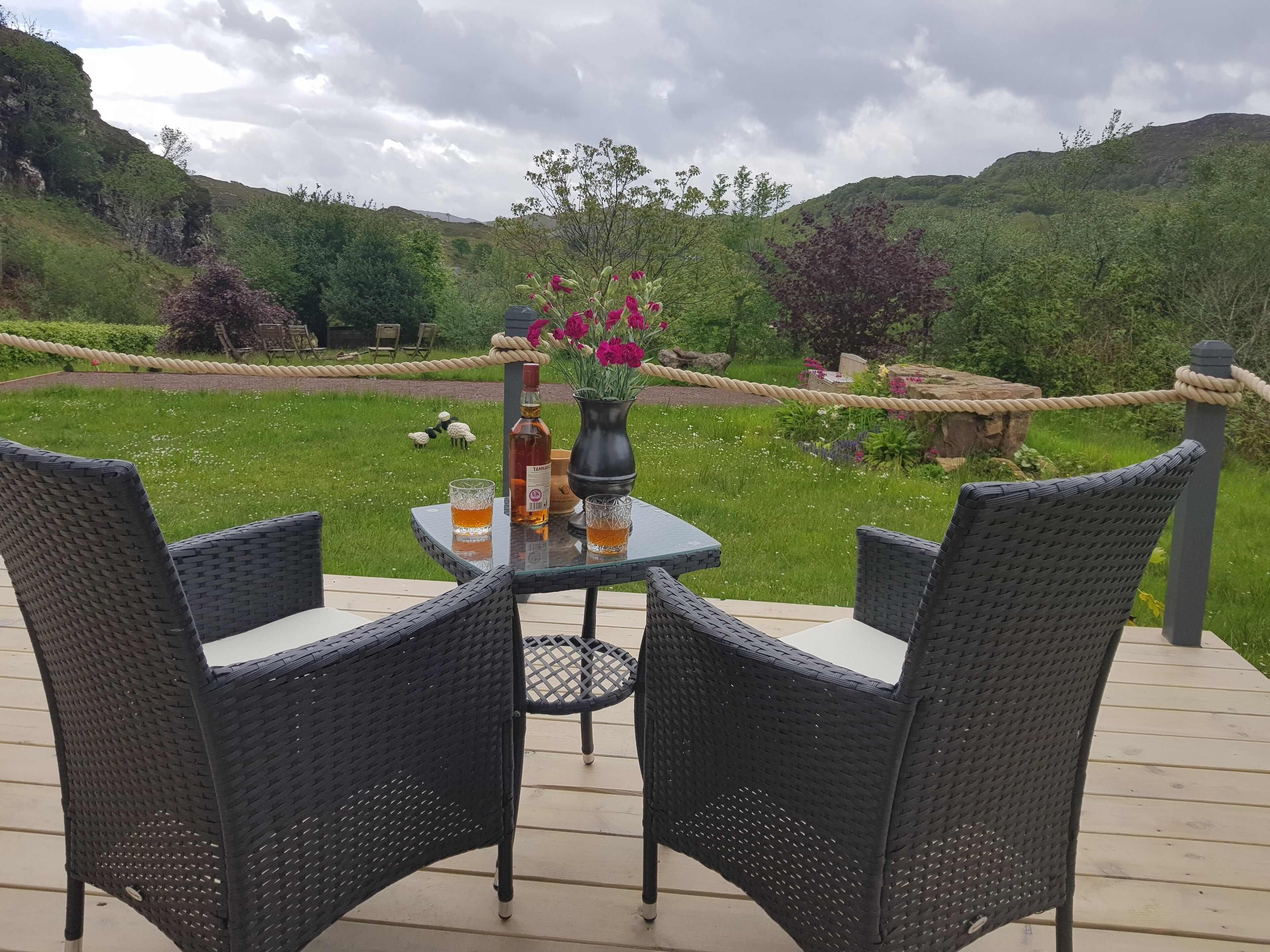 A deck with wicker chairs and a table, overlooking a lush garden with a rope barrier.