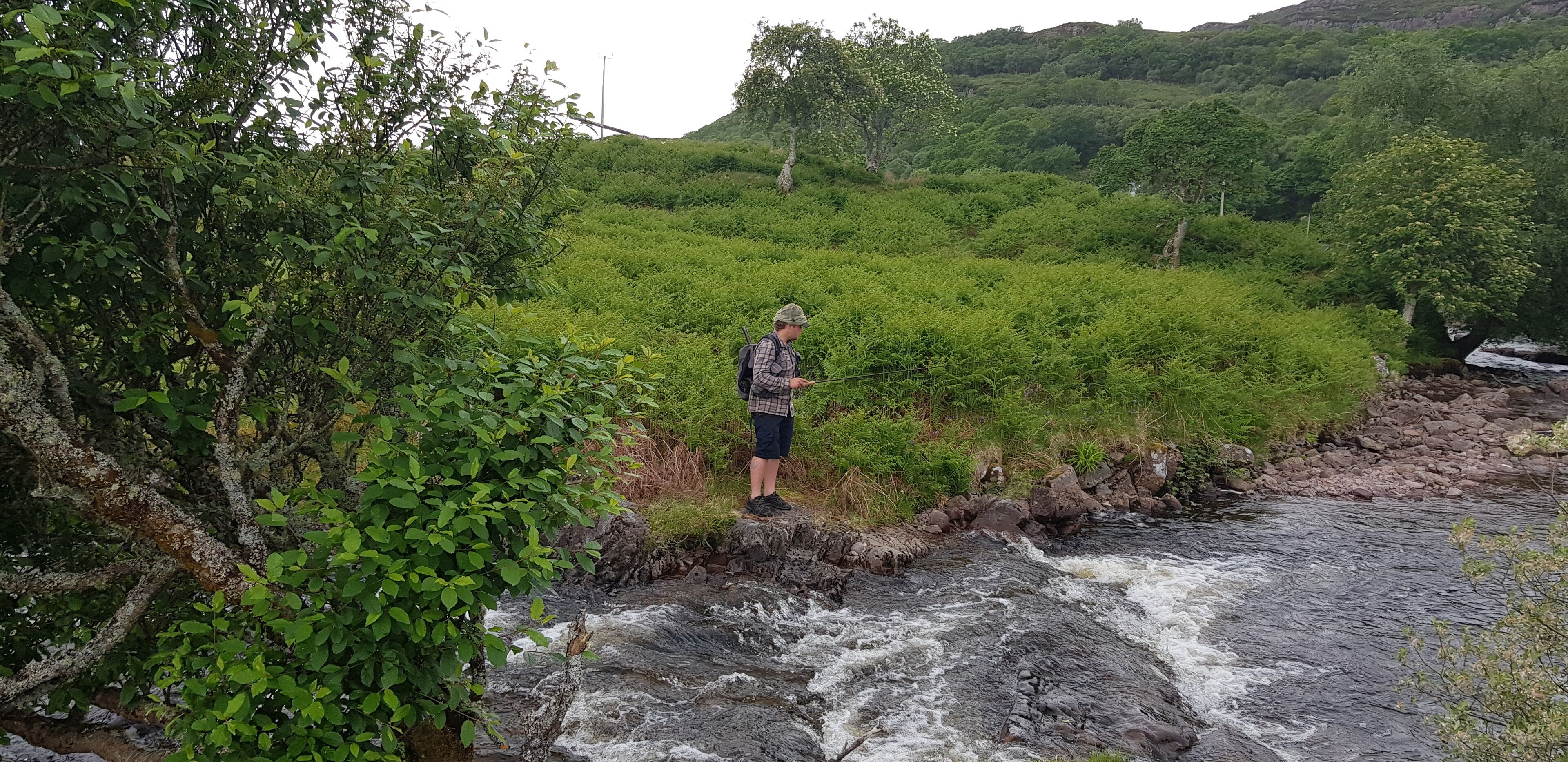 Una persona está de pie junto a un río caudaloso en una zona exuberante y verde, con vegetación densa y colinas al fondo.