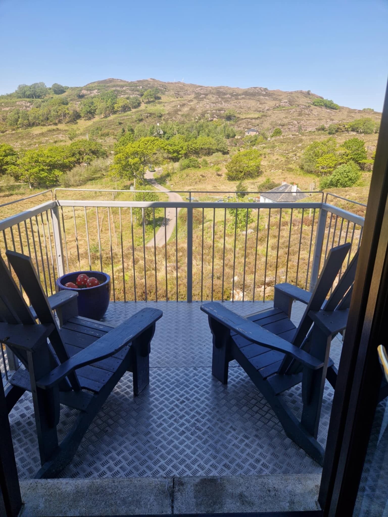 Ein Balkon mit zwei Holzstühlen und einer Obstschale. Der Balkon bietet Blick auf eine hügelige Landschaft mit Bäumen und einer Straße.