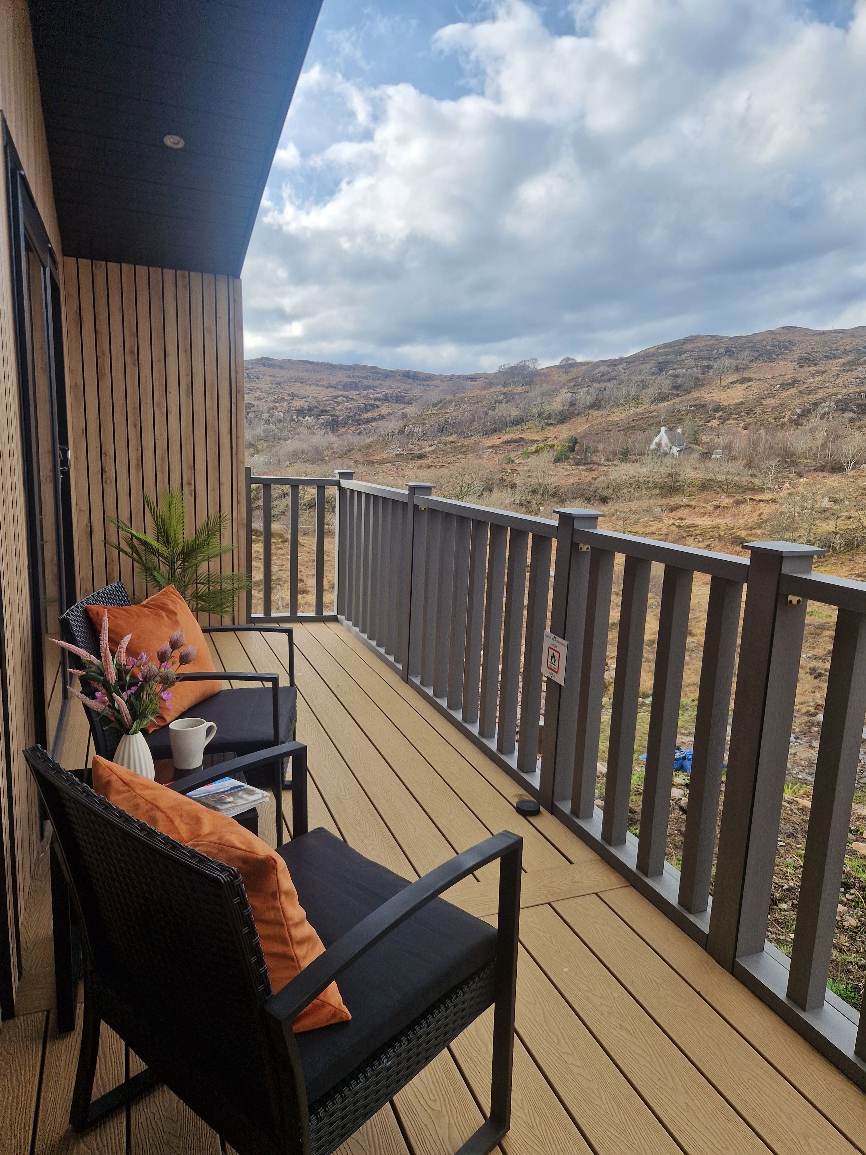 Una terraza de madera con mobiliario exterior, vista a un paisaje montañoso y cielo nublado.