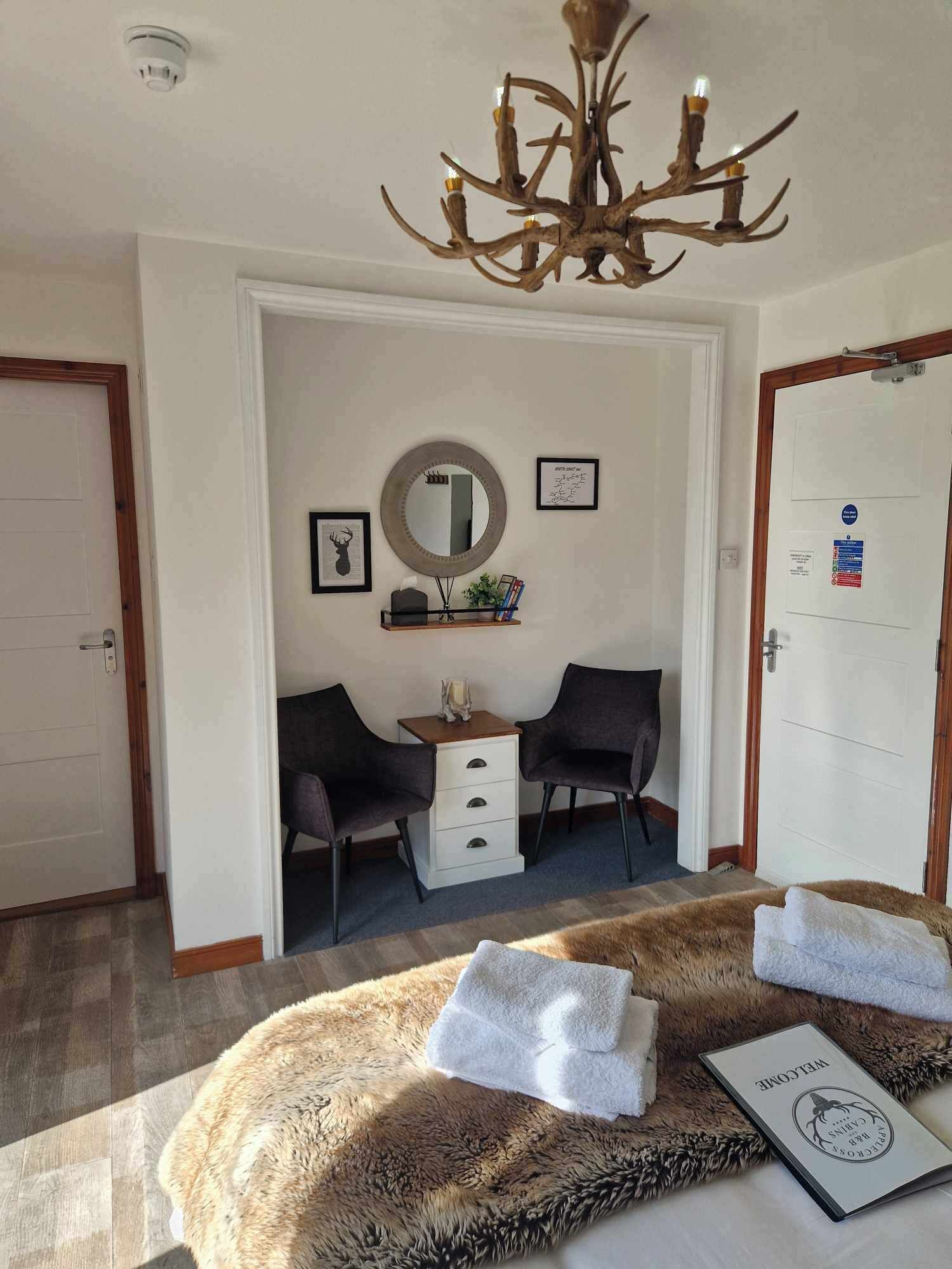 A hotel room with a chandelier, a mirror, two chairs, a dresser, and a fur rug with towels.