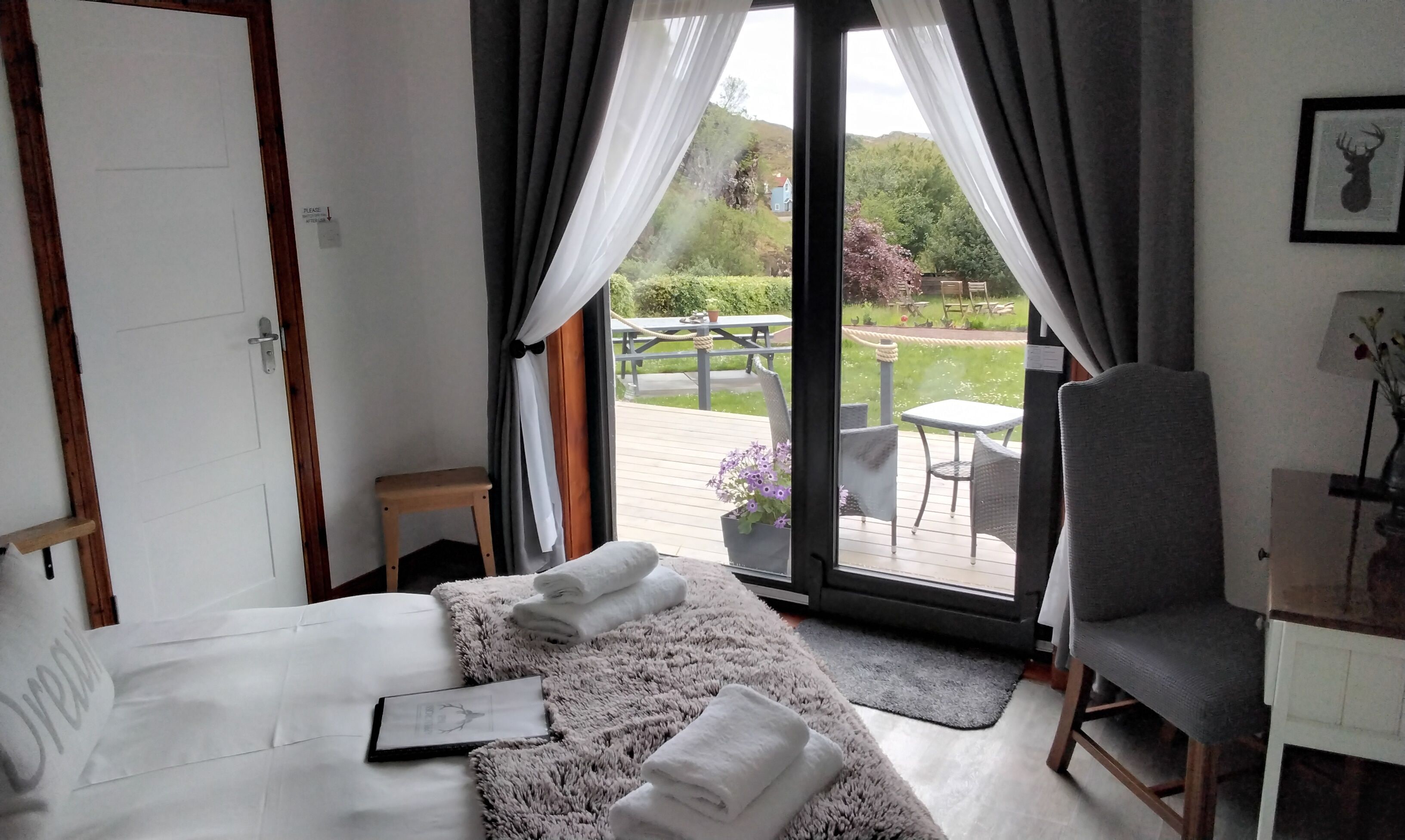 A bedroom with a bed, a chair, a small table, and a view of an outdoor deck with a table and chairs.
