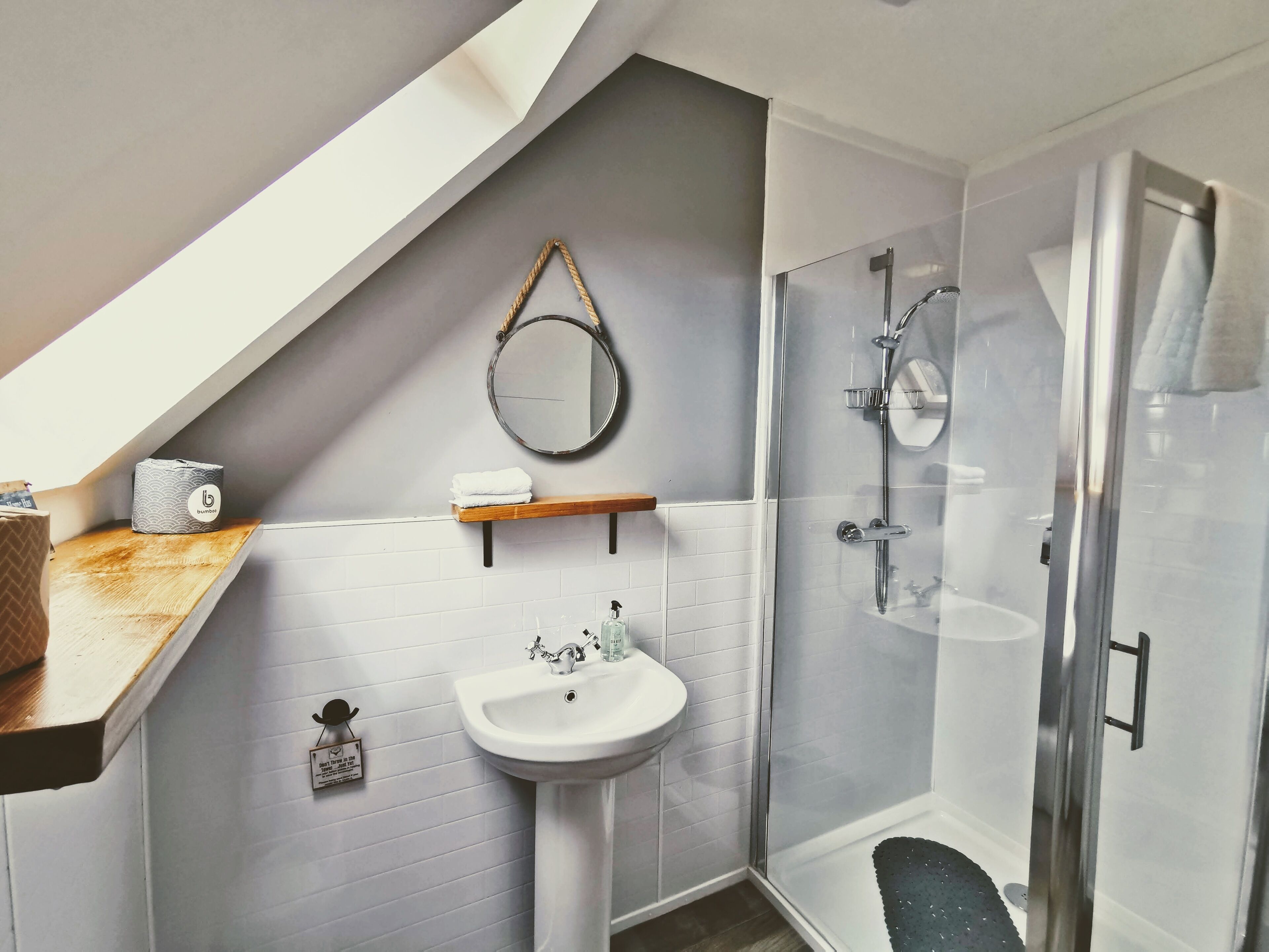 A modern bathroom with a white sink, a glass shower enclosure, and a wooden shelf with a towel and toilet paper.