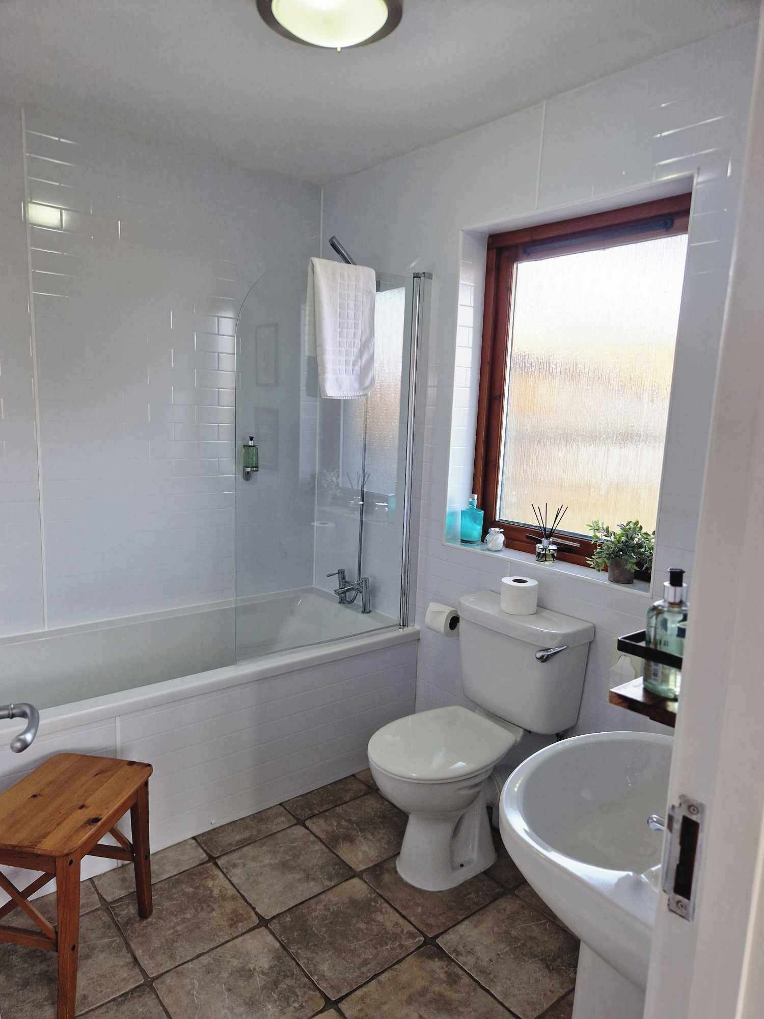 A bathroom with a glass-enclosed shower, a toilet, a sink, and a wooden stool.