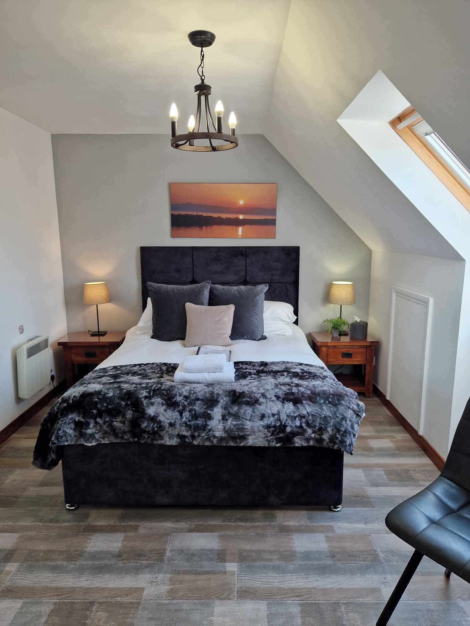A bedroom with a large bed, a chandelier, a painting of a sunset, and a skylight.