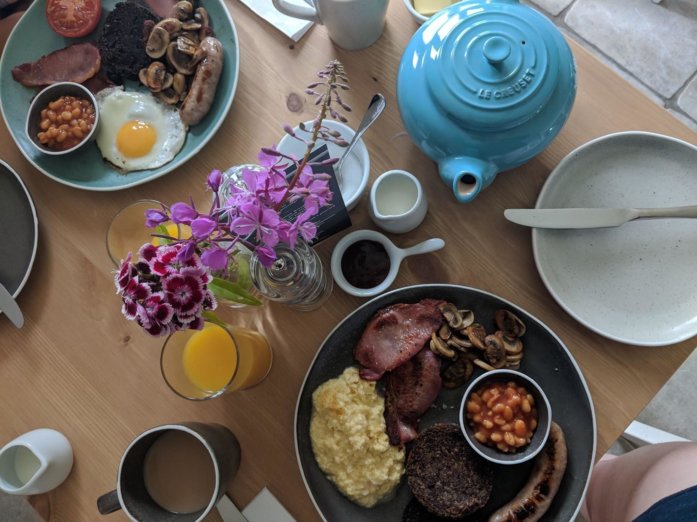 A breakfast table with plates of eggs, sausages, beans, and ham, a blue teapot, and a glass of orange juice.