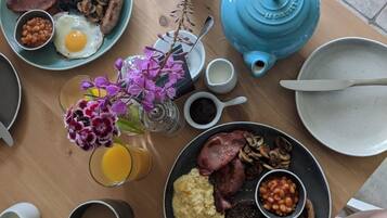 A breakfast table with plates of eggs, sausages, beans, and ham, a blue teapot, and a glass of orange juice.