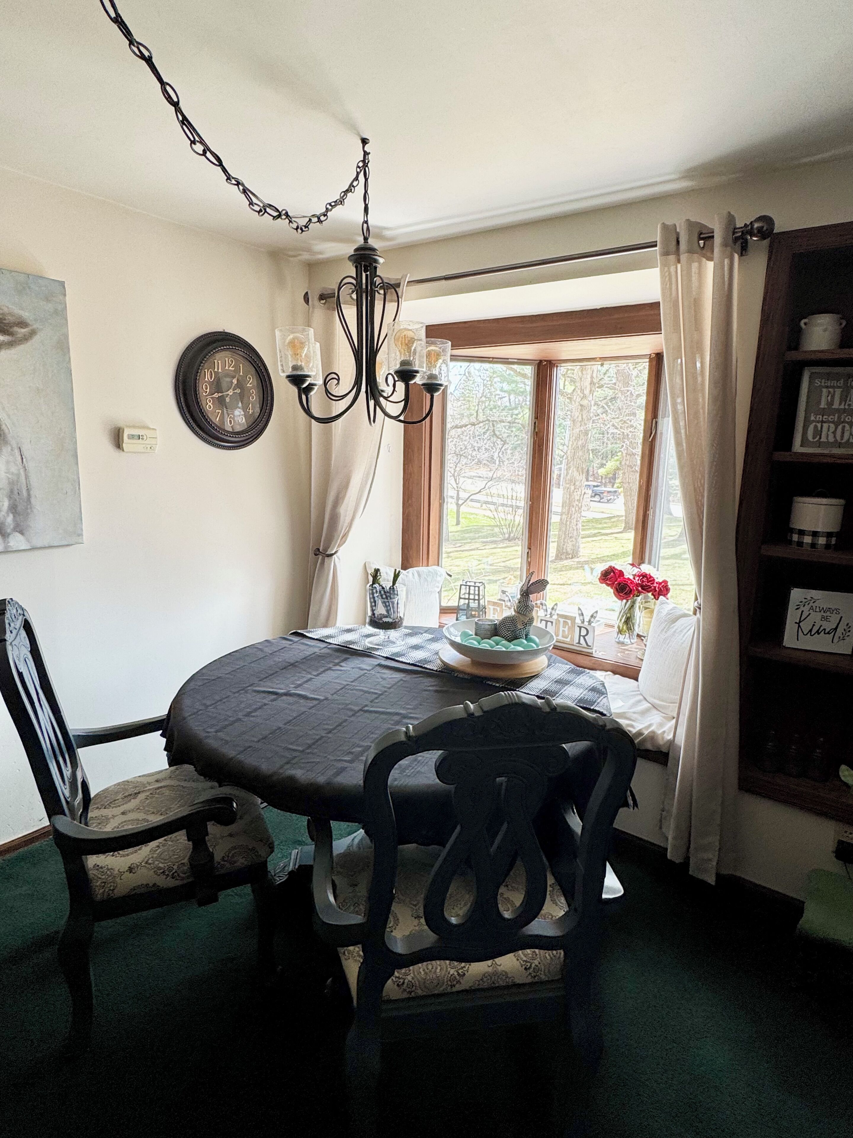 A dining area with a round table, chairs, a chandelier, a window with curtains, and a bookshelf.