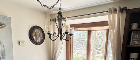 A dining area with a round table, chairs, a chandelier, a window with curtains, and a bookshelf.