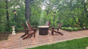 Un patio con una mesa redonda y cuatro sillas Adirondack, rodeado de exuberante follaje y un camino de ladrillo.