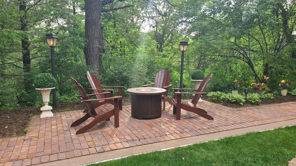 Un espace patio avec une table ronde et quatre chaises Adirondack, entouré d’une végétation luxuriante et bordé d’un chemin en briques.