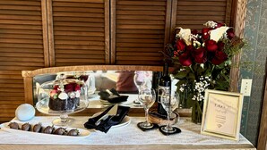 Una mesa dispuesta con un pastel bajo una campana de cristal, fresas bañadas en chocolate, copas de vino, un ramo de rosas rojas y un cartel enmarcado.
